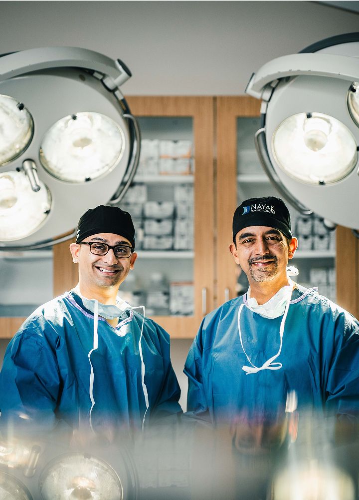 Top St. Louis facial plastic surgeon Dr. Nayak and Dr. Kachare smiling in an operating room setting.